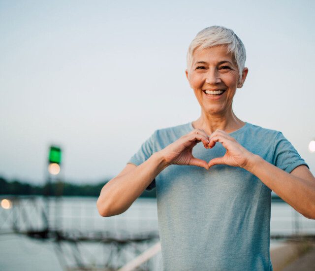 elder woman standing next to a lake making the heart sign with her hands