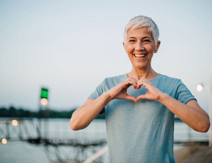elder woman standing next to a lake making the heart sign with her hands