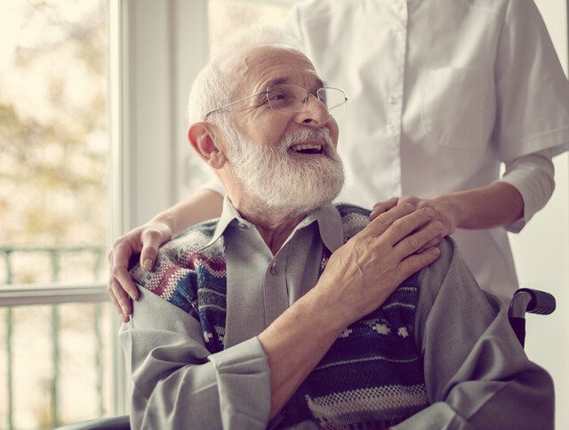 Senior man sitting on the wheelchair, laughing and holding his nurse's hand