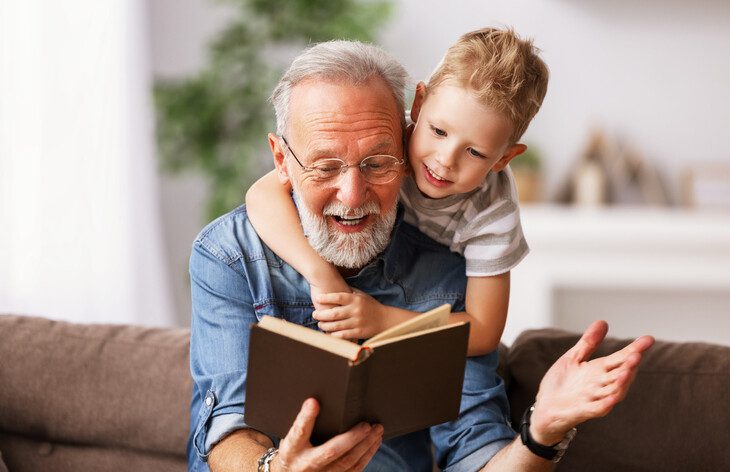 Happy family elderly man and little boy smiling while sitting on couch and reading fascinating fairy tale together at home