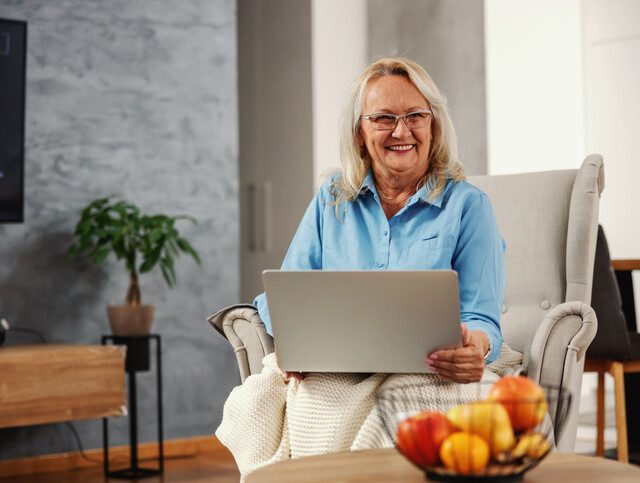 Smiling senior woman sitting at home in her chair with laptop in her lap and hanging on internet.