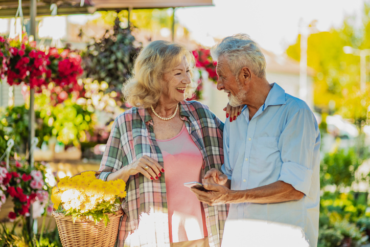 A senior couple is on the market, a senior woman is carrying flowers and smiling and a senior man is holding a mobile phone