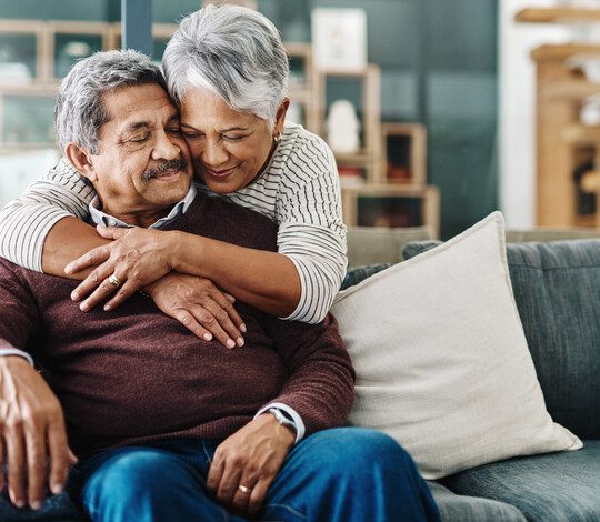 woman hugging her husband from behind as he's sitting on the couch