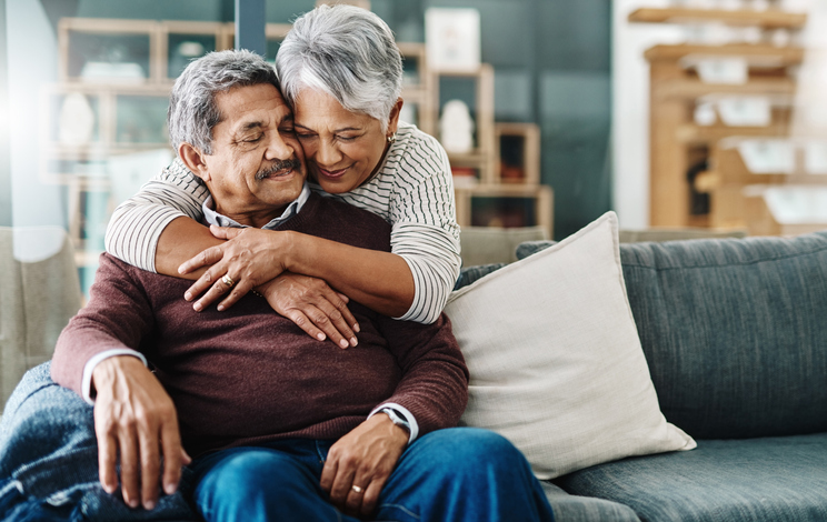 woman hugging her husband from behind as he's sitting on the couch