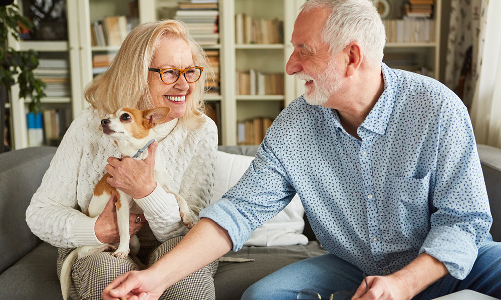 smiling senior couple sitting on a couch smiling with their dog.